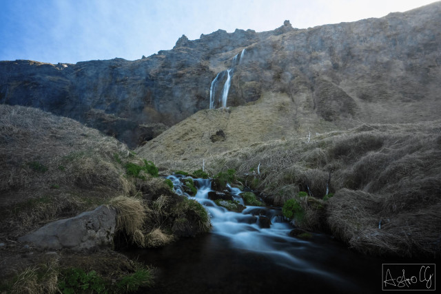 Mountain landscape with waterfall flowing over rocks into a small stream surrounded by grass and rocks under a blue sky