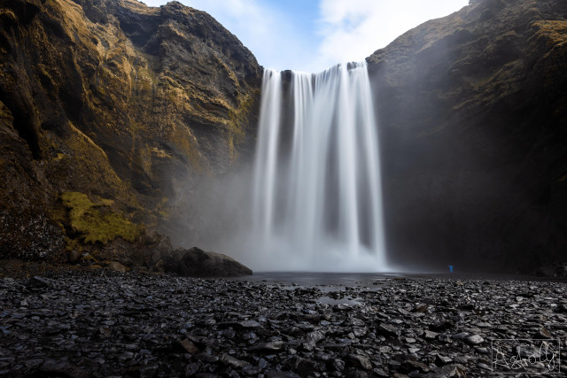 Waterfall cascading from a high cliff into a river with rocks and pebbles in the foreground