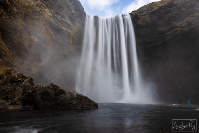 Waterfall cascading from a high cliff into a river with rocks in the foreground