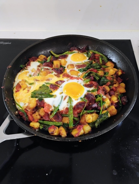 Frying pan on a black glass hob. It contains baked eggs in a mixture of tomatoes, chillies, chorizo, fried potatoes, spinach, onion and garlic. Fabulous contrast of red, green, white and yellow.