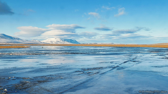 Four colour photos of snow scenes. This one is of a wide expanse of water which has frozen. There are pressure ridges and cracks in the ice, suggesting that there might be flow beneath the ice. A strip of golden pasture land is visible on the other side. In the distance is a range of snow-clad mountains with fluffy white cloud above them. The rest of the sky is clear cyan, and this is reflected in the ice.