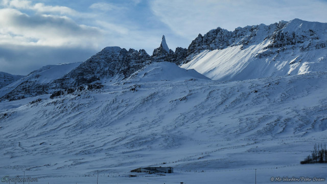 A mountainside covered in snow and ice, with farm buildings near the foot. The tall mountain is ridged and eroded, these contours are highlighted by the snow. In the centre at the top is a lovely pointed spire of rock, surrounded by other peaks forming a ridge along the apex. The sunlight from the left is catching some, but not all, of the contours.