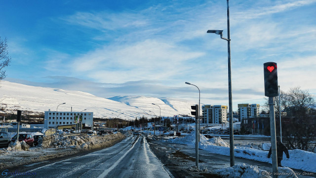 A road into a large town. Piles of snow are heaped beside the tarmac and pavements have been cleared. Streetlights are lining the route and in the distance is an impressive mountain range, white-coated with snow and ice. Near to the viewpoint is a traffic signal, showing red, and the light has been painted into a red heart. The sky is mostly clear and the sunlight is from the right.