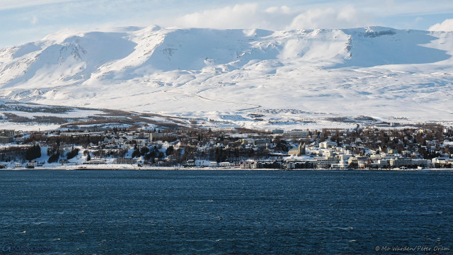 Finally, a shot of a large town on the opposite side of a body of water. The surface of the fjord is ruffled by wind and is blue in reflection of the sky. Sunlight is from the left. Right from the waterline, the town is busy with buildings and infrastructure. Right of centre is an impressive twin-spired church. There are residences and other structures as far away as the foothills of a large mountain. A road can be seen climbing the mountain towards the ski slopes.