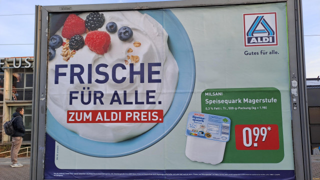Aldi-Werbeplakat für Quark. Als Produktabbildung ist ein hellblauer Teller mit Quark dargestellt, auf dem ein paar Heidelbeeren, Himbeeren und Haferflocken drapiert sind.