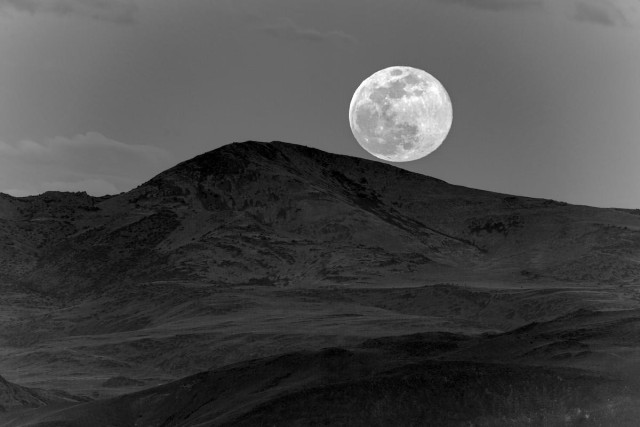 A black and white photo showing a full Moon rising over a hill. The Moon almost looks like it is rolling down the hill to the right. Textures are seen in the dark hillside. The sky is a dark gray with a few clouds. The Moon is big and bright showing its textures of light and dark places on the surface.