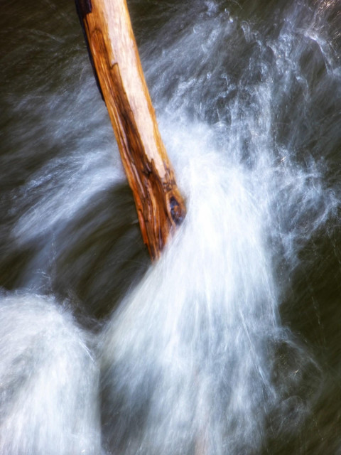 A large branch sticks out of the water in a fast moving stream. Water splashes and twists around the branch and the colours and softness of the water gives the photograph a painting-like look.