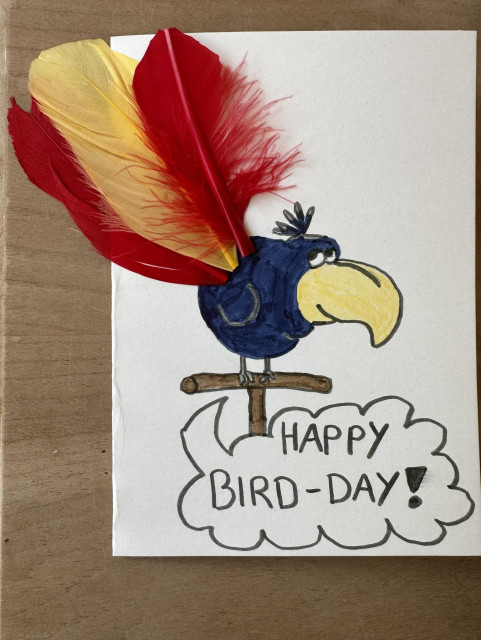 A colorful birthday day card featuring a blue cartoon bird with a yellow beak, perched on a branch. The bird has red and yellow feathers attached to the top and includes the message "HAPPY BIRD-DAY!" in a speech bubble. Yes, it’s my birthday 🥳