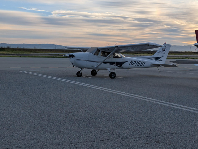 A Cessna 172S high-wing airplane sitting on the ramp at San Carlos. The San Fransciso east bay hills are far in the background. There are high clouds, which are orange from the rising sun.