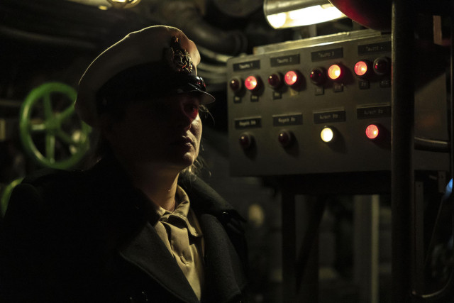 Eine weiblich gelesene Person mit Offiziersmütze in einem U-Boot. Ernster Blick. Im Hintergrund rote Signallampen an einem Diesselpunk-ähnlichem Kontrollkasten sowie ein grün angestrahltes Ventil-Rad.