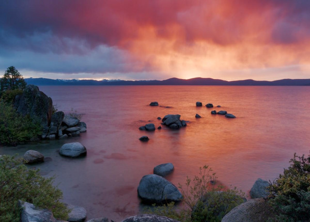 A color landscape photo of a sunset over a large lake. The photo is shot from a small cove surrounded by large gray boulders and green shrubs. A small conifer tree is seen on the far left. A series of boulders extend out into the lake from the lower central part to about center frame. In the far distance across the large lake is a low range of mountains. The sky is cloudy with dark rain clouds. But the central part of the clouds is bright orange and red from the setting sun. The sunset colors are also reflected in the lake water where the boulders are near center frame. The lake is flat and it is a very tranquil yet colorful scene.