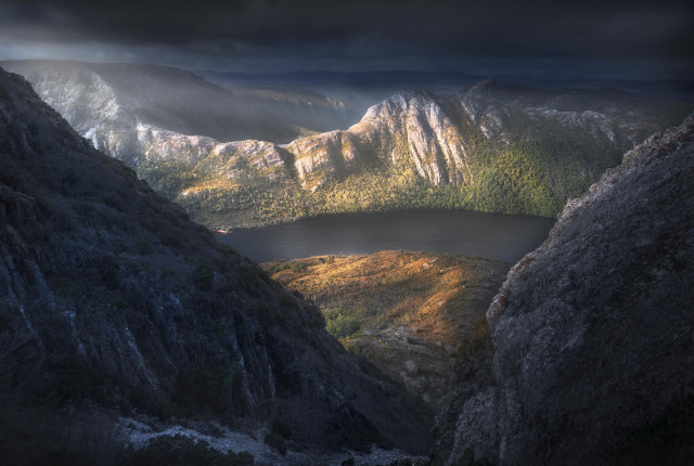 Moody mountain landscape with dark storm clouds, sunlit cliffs and forest surrounding a calm alpine lake, viewed from a rocky ridge.
