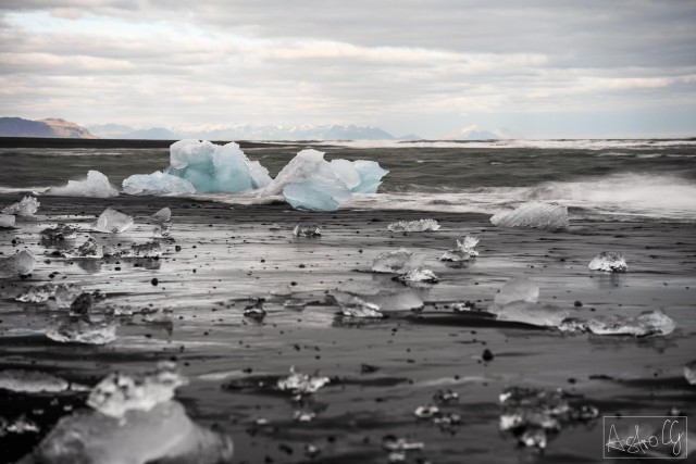 Black sand beach with scattered ice chunks and a larger ice block in the water under a cloudy sky