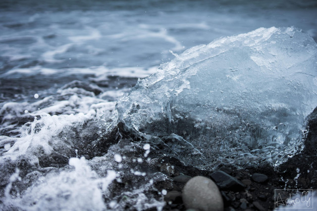 A large piece of ice lies on the shore surrounded by waves and small stones