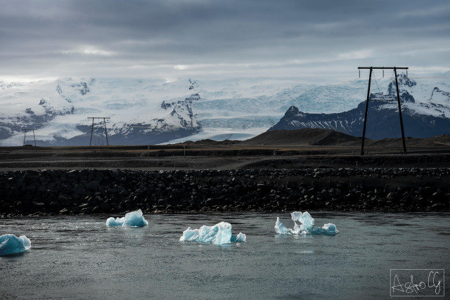 River with floating ice chunks in front of a barren landscape with power poles and snow-covered mountains in the background