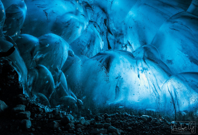 View from inside a blue-lit ice cave with smooth, curved ice surfaces and a rocky floor