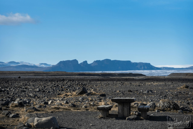 Rocky barren landscape with a simple wooden table and two benches in the foreground and a flat mountain range in the background under a blue sky