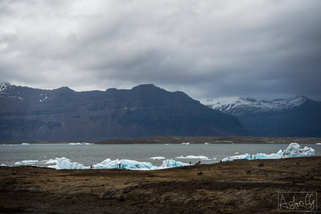 Landscape with dark mountains, snow-covered peaks, water with floating ice chunks, and cloudy sky