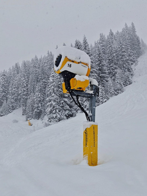 A photo of a yellow snow-covered snow cannon on a snowy ski slope with snowy trees in the background.