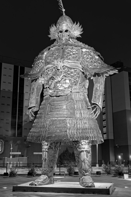 A black and white portrait photo of a large statue made of metal. It is of a skeleton wearing ancient armor like the armor of Genghis Khan. It is filled with Asian motifs and designs. It is night so the statue is lit from the right. In the background is a hotel / casino building.