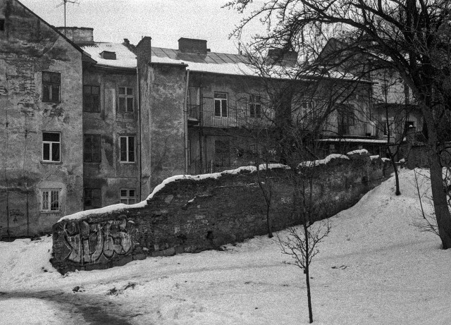 A black-and-white photograph of an old residential courtyard in one of the historic central districts of Lviv. Weathered buildings with peeling plaster and uneven facades stand close together, their windows mismatched and timeworn. A narrow balcony with a simple metal railing runs along the second floor, hinting at quiet domestic life behind the walls.

In the foreground, a crumbling stone wall cuts diagonally through the frame, its rough surface partially covered with graffiti. Snow rests heavily on top of the wall and blankets the sloping ground below, softening the harsh textures of brick and concrete. Bare trees rise from the snow, their thin branches reaching upward against a pale winter sky.

The scene feels still and subdued — a fragile balance between decay and endurance, where history lingers in every cracked surface and silent window.
