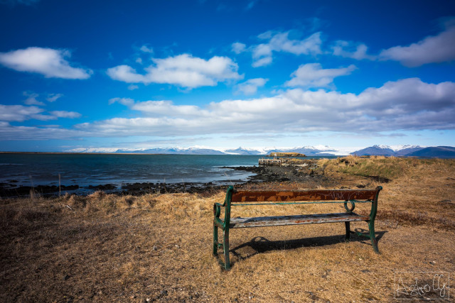 Empty park bench on dry grass overlooking the sea and snow-covered mountains under a cloudy sky