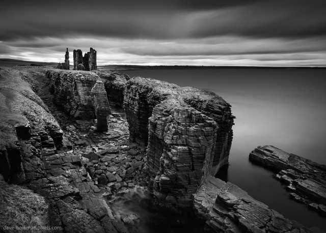 A photograph of Castle Sinclair Girnigoe, an ancient ruined castle standing on the edge of tall cliffs above the rugged coastline of Caithness in Scotland. The structure rises directly from the rock, its broken walls and empty window openings clearly defined against a heavy, dark sky. The long exposure smooths the movement of the sea below, so the water appears misty and soft around the base of the cliffs. Overhead, thick clouds create a brooding atmosphere, making the castle feel isolated and imposing against the wild coastal landscape.