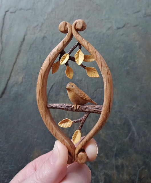 A small wood carving of a tiny bird in golden brown perched on a branch, looking from right to left. There is a small branch protruding from that branch with two carved leaves. Above the bird are two further branches, one behind with four leaves, and one in front with two leaves. All of it is held between two scrolls of light wood, that form an oval around it. Held at the bottom by a finger and thumb. With a grey slate background.