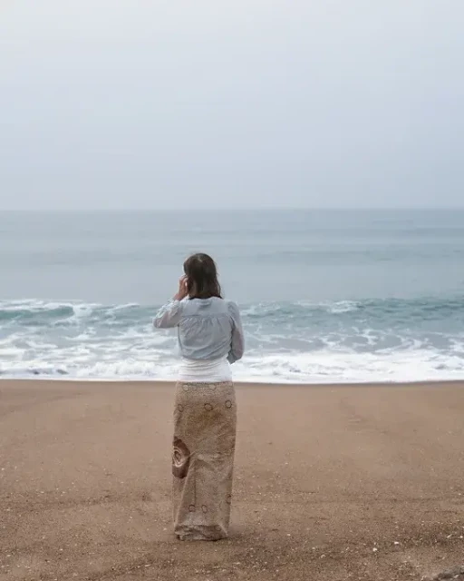 A staged photo as a woman stands on a sandy beach looking out to sea. She blends perfectly with her skirt matching the sand, a waistband matching the white surf and her top matching the blue-grey of the sea