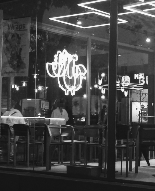 Black and white photo of a restaurant window with a neon logo that's reflected in the glass and looks like a pair of cartoon boobs.