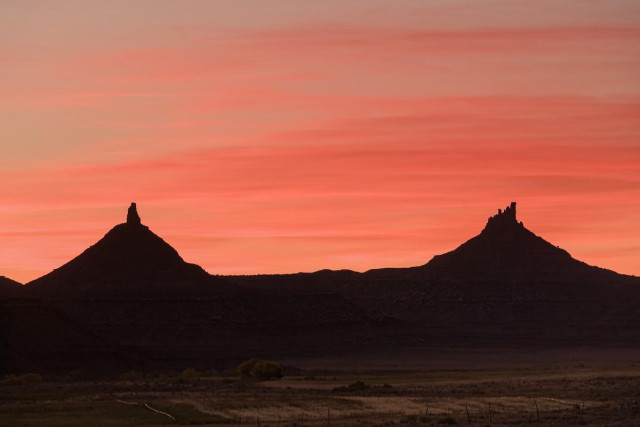 A color landscape photo of a pair of eroded sandstone peaks. There is one of the left and one on the right. They are very nearly the same in height and look. Each has a conical base with a thin protruding top giving them the appearance of pistols pointing toward the sky. In the foreground is tilled fields that are deep in shadow. The sky is striped with red, pink, and orange sunset colors. 