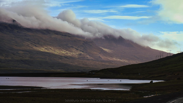 A colour photo of the edge of a body of shallow water. The black sands and surrounding rock is all in shadow from the mountain behind us. Ahead is a mountain range, its flanks a patchwork of shade and sunlight from the right. The sky is mostly clear cyan but the pinnacles of the mountain range are capped by dense cloud.