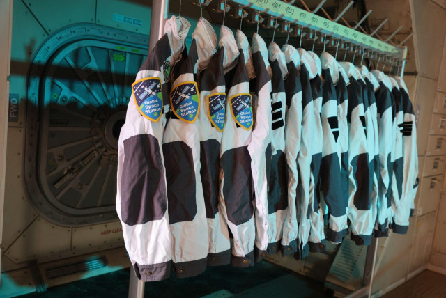 Eine Reihe von weiß/anthrazit-farbingen Jacken auf einem Kleiderständer vor einem vage Luftschleuse-ähnlichem Kulissenelement. Auf einigen der Jacken sind Ärmel-Ptches mit "Global Space Station". 