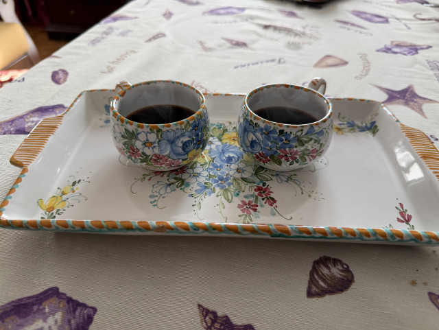 Two small floral espresso cups filled with coffee sit side by side on a matching hand-painted tray, placed on a tablecloth with seashell patterns. Light steam rises from the cups, creating a quiet, intimate moment.

