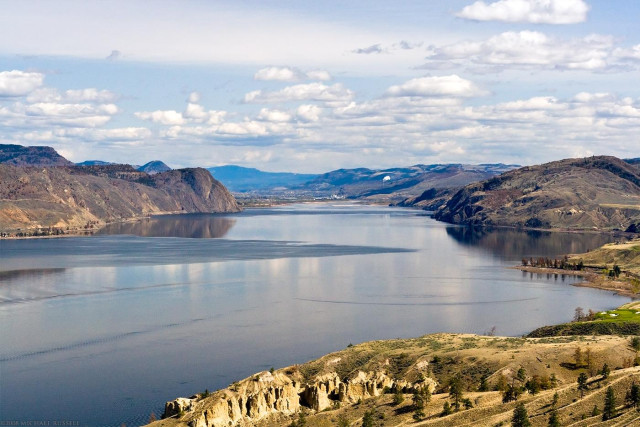 View of a large lake with sagebrush areas on the surrounding hills, and the city of Kamloops at the other end of the lake, on mostly clear day with spotty clouds in the sky.