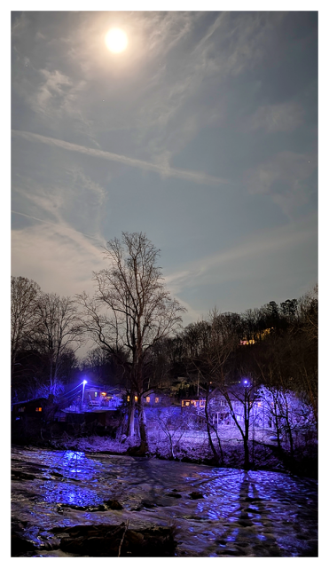 a full-appearing moon illuminates a night scene with leafless trees by a gently flowing river. purple-blue lights cast an ethereal glow on the water and trees, while a few small houses with lit windows are visible in the background under a cloudy sky.