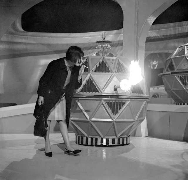 A woman in fashionable 1960s attire standing on a futuristic city set. She is next to a large spherical robot with a flame thrower sticking out of it. The flame thrower is shooting out a metre of flame and the woman is leaning casually in to light a cigarette.