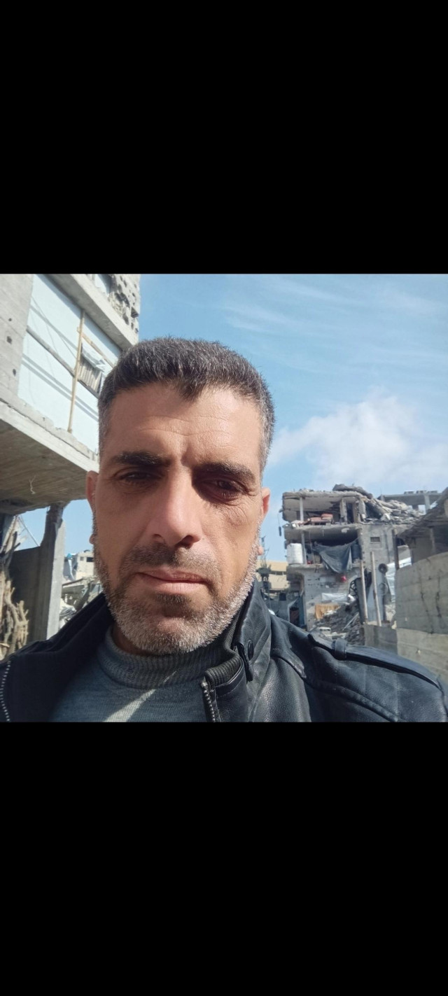 "Close-up of a middle-aged man with dark, graying hair and a short beard, looking directly at the camera with a serious expression. He is wearing a gray turtleneck sweater and a dark jacket. Behind him, under a clear blue sky, there are severely damaged and ruined buildings with scattered debris, suggesting an area affected by conflict or disaster."