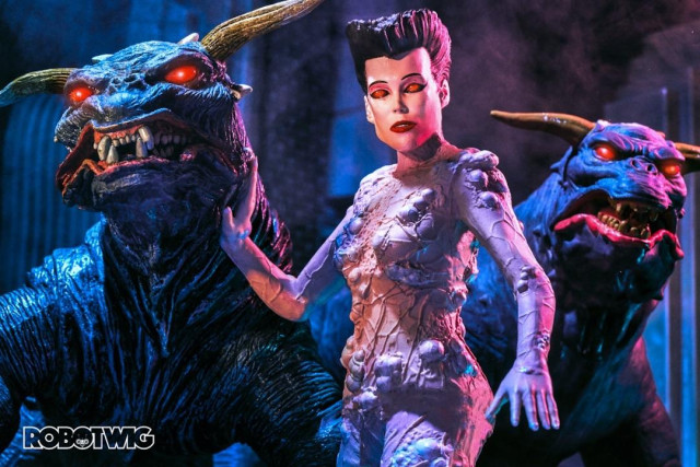 Gozer looks into the camera with glowing red eyes, she strokes one of the terror dogs whos eyes are also glowing red, another dog looks at the camera behind her