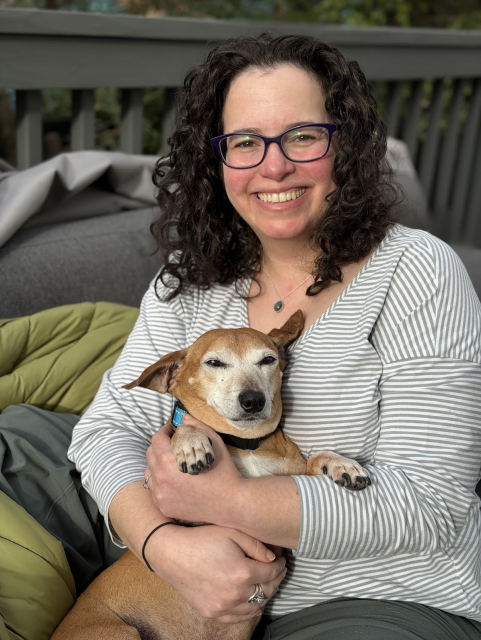 A smiling white woman with dark curly hair is sitting outside on a sunny day, holding a dachshund-mix upright with his belly towards the camera, in her lap. The dog is squinting and not really sure why this is happening to him, he doesn't deserve this. 