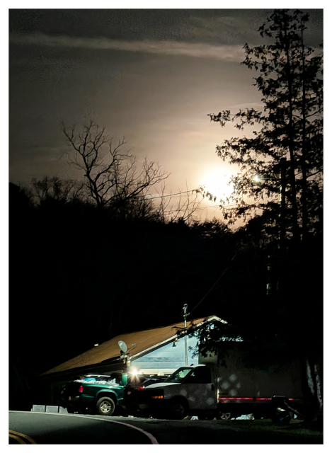 house with a slanted roof is illuminated by exterior lights at night. a truck and trailer are parked outside. leafless trees and a tall evergreen stand nearby, with a bright full moon glowing in the cloudy night sky behind them.
