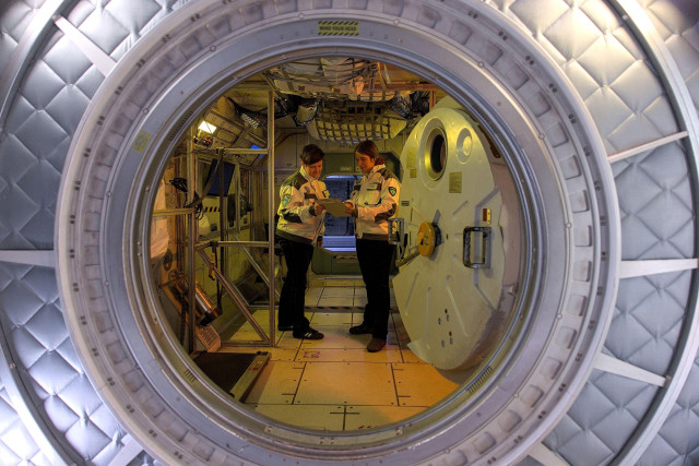 Zwei Frauen in einer Raumstation-Kulisse, fotografiert durch eine runde Luftschleuse. Sie tragen weiße Uniformjacken mit Mission-Patches und diskutieren über einem Clipboard.
