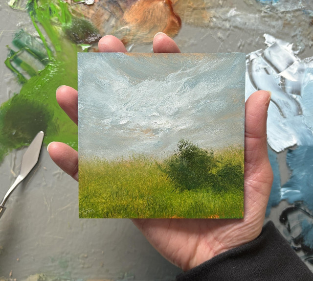 Photo of a hand holding an original landscape oil painting by Tisha Mark, "Homeland No. 2603" oil on Ampersand Gessobord, 4"x4" (2026). A peaceful atmospheric landscape painting of a lush green meadow with a couple loosely painted trees on the right. There are undertones of orange in the blue-gray sky and in the grass. A paint palette with puddles of oil paint and a palette knife are visible in the background.
