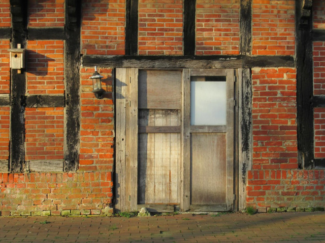 Eine Tür in einer alten Fachwerkfassade aus rotem Klinker und Balken, von denen die schwarze Farbe so langsam verschwindet. Es ist keine "richtige" Tür, sondern eher ein zusammengewürfeltes Provisorium aus unbehandeltem Holz. Rechts davon eine Lampe und eine Nisthilfe.