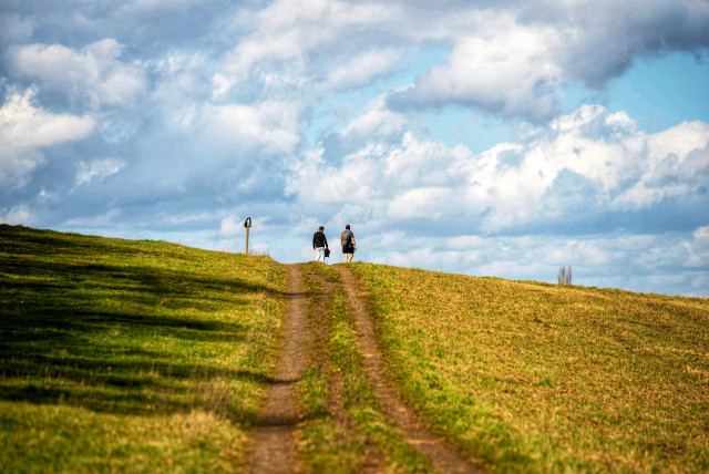 Das Bild zeigt eine Landschaftsaufnahme, die sich auf einen grasbewachsenen Hügel konzentriert, der von unten nach oben fotografiert wurde. Im Vordergrund dominiert ein Feldweg, der sich durch grünes und gelbes Gras schlängelt und zum oberen Rand des Hügels führt. Der Blickwinkel ist so gewählt, dass der Hügel fast die gesamte untere Hälfte des Bildes einnimmt. Am oberen Rand des Hügels, vor dem Horizont, sind zwei Personen zu sehen, die in die Ferne blicken oder weitergehen. Die Person links trägt dunkle Kleidung und eine helle Tasche, die Person rechts trägt ebenfalls dunkle Kleidung. Links neben den Personen steht ein kleiner, dunkler Pfahl. Der Himmel im oberen Teil des Bildes ist dynamisch und mit dramatischen, bauschigen weißen und grauen Wolken gefüllt, die einen Kontrast zum hellblauen Himmel bilden. Die Beleuchtung ist hell, mit deutlichen Schatten auf der linken Seite des Hügels, was auf Sonnenlicht von rechts oben hinweist.