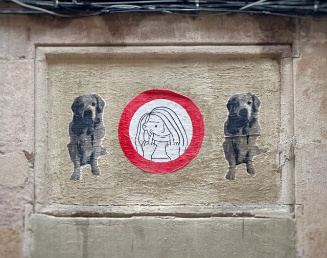 Gatukonst på en husvägg. I mitten en varningsskylt med ett tecknat kvinnoansikte som håller upp sina mungipor i ett leende med två fuck-you-fingrar, bredvid två svartvita hundar.