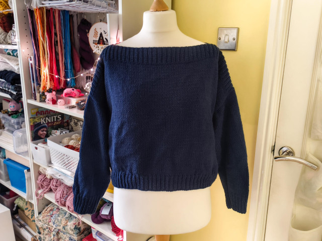 A navy blue jumper on a dressmakers dummy. It has a wide ribbed boatneck and is otherwise plain stockinette, with long sleeves. In the background, many colourful and disorganised craft supplies on various shelves.