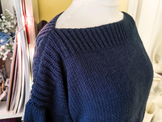 Angled close-up on the neckline and shoulder of the same navy jumper. The ribbing of the neckline contains down the shoulder for a few inches before turning into stockinette sleeve.