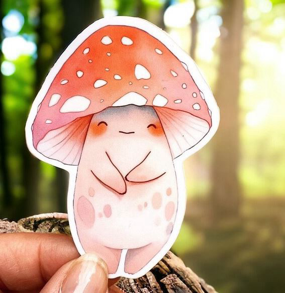 Adorable watercolor illustrated mushroom character sticker with a red and white spotted cap and a shy smiling face. Fingertips hold the sticker in front of a sunlit forest, creating a whimsical woodland feel.