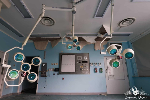 An eerie abandoned operating room features multiple surgical lights with green and blue lenses, a central control panel, and a damaged ceiling